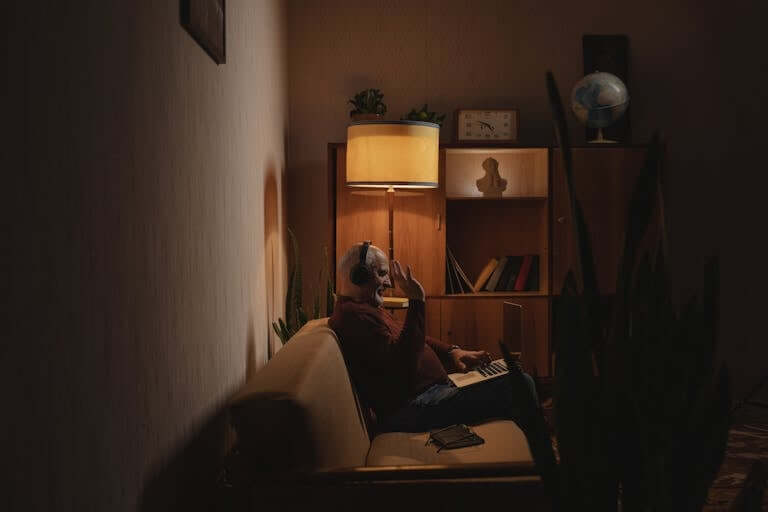 An elderly man sitting on a couch indoors, using a laptop for a video call at night.